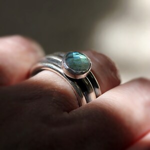 May include: Close-up of a silver ring featuring a round, pale blue gemstone. The ring is on a finger, with other silver bands visible. The gemstone has a faceted surface, reflecting light.