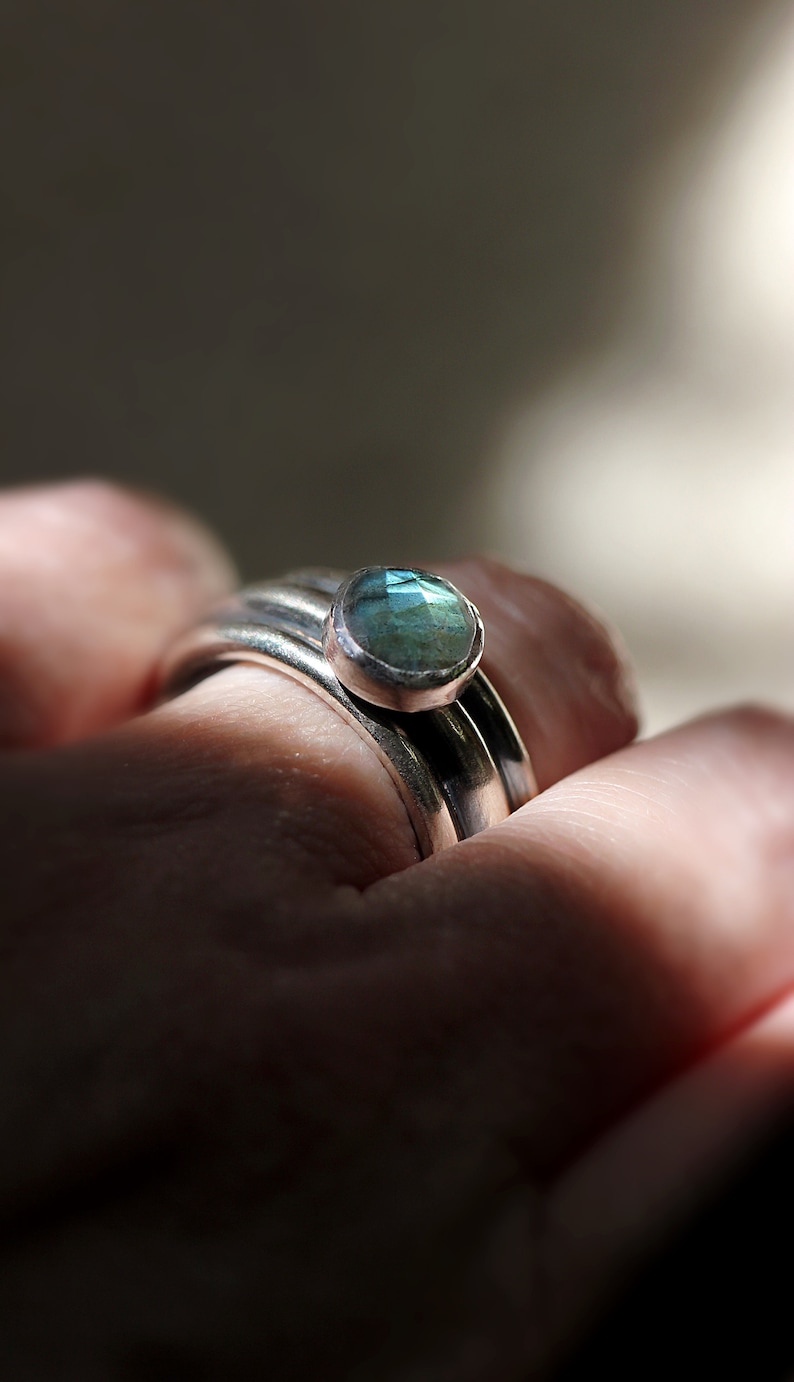 May include: Close-up of a silver ring featuring a round, pale blue gemstone. The ring is on a finger, with other silver bands visible. The gemstone has a faceted surface, reflecting light.