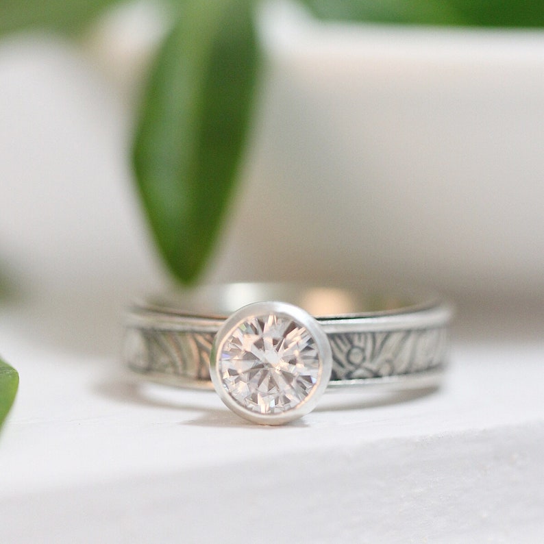 May include: A silver ring with a large, clear, round gemstone. The band features an engraved pattern. The ring is set against a white background with a blurred green leaf in the background. Jewelry.