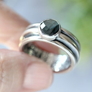May include: A silver ring featuring a dark, faceted gemstone. The ring has a multi-band design, with the gemstone set atop the band. The ring is held by a person's finger.