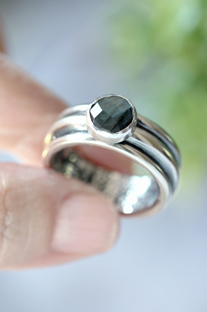 May include: A silver ring featuring a dark, faceted gemstone. The ring has a multi-band design, with the gemstone set atop the band. The ring is held by a person's finger.