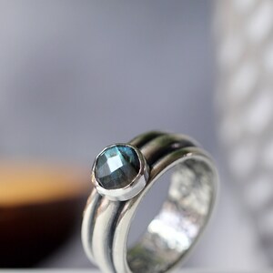 May include: A silver ring with a round, blue-green gemstone. The ring has a textured band with three ridges. The gemstone is set in a smooth bezel. The ring's design is elegant and eye-catching, suitable for everyday wear.