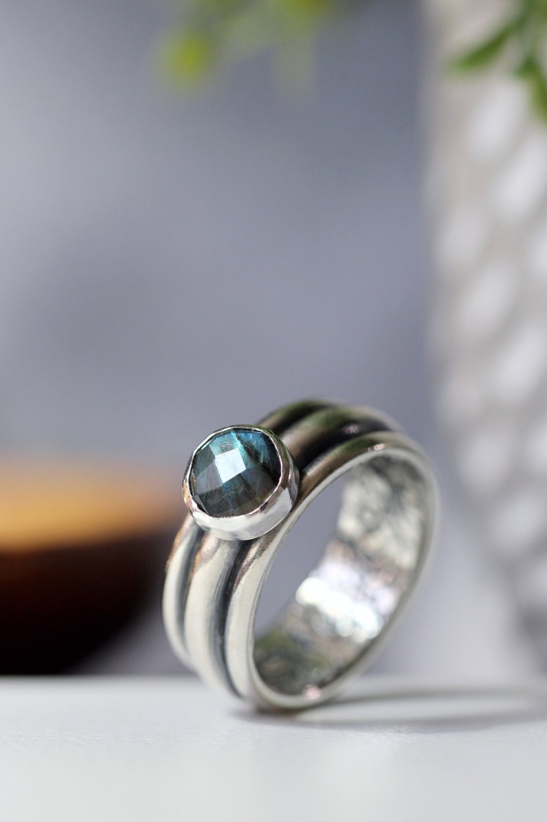 May include: A silver ring with a round, blue-green gemstone. The ring has a textured band with three ridges. The gemstone is set in a smooth bezel. The ring's design is elegant and eye-catching, suitable for everyday wear.
