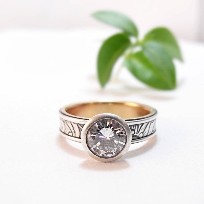 May include: A silver ring with a large, clear gemstone set in a bezel. The band features an engraved floral pattern. The ring has a gold interior and is set against a white background with a blurred green plant in the background.