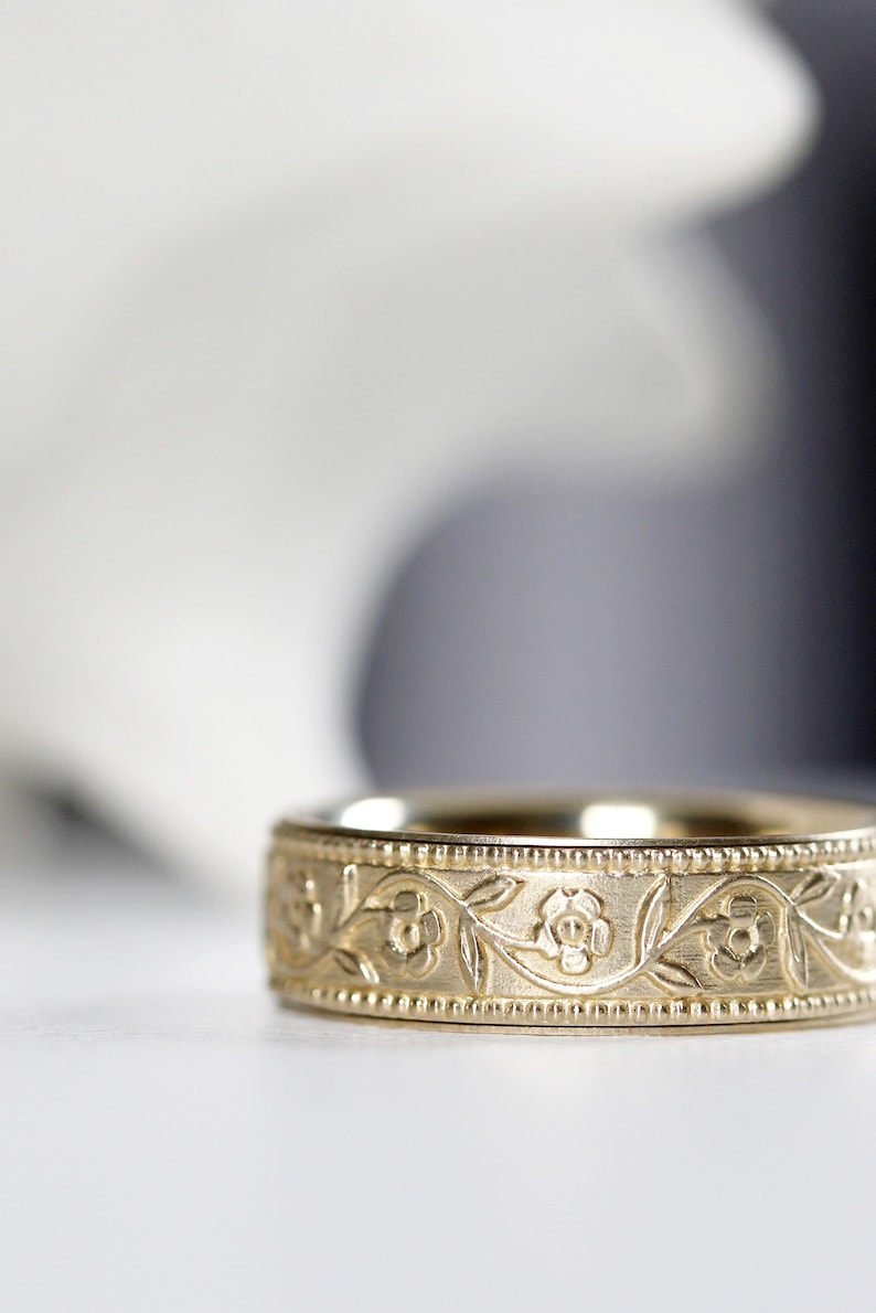 Op de afbeelding: Een gouden trouwring met een bloemmotief. De ring is gegraveerd met een delicaat patroon van bladeren en bloemen.