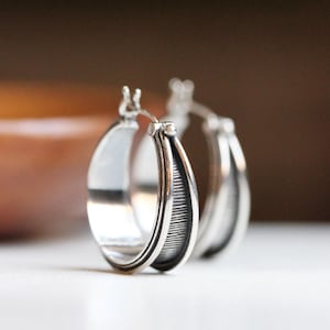 May include: A pair of silver hoop earrings featuring a wide, polished outer band and a recessed, dark, vertically ribbed inner section. The earrings have a hinged post closure mechanism visible at the top of each circular hoop design.