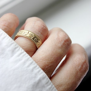 Op de afbeelding: Een gouden ring met een bloemmotief. De ring wordt aan een vinger gedragen.