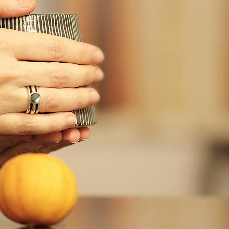 May include: A black and white striped ceramic mug held by a person wearing a silver ring with a blue-green stone. A small, orange pumpkin sits in the foreground. The mug has vertical stripes and a slightly flared shape.