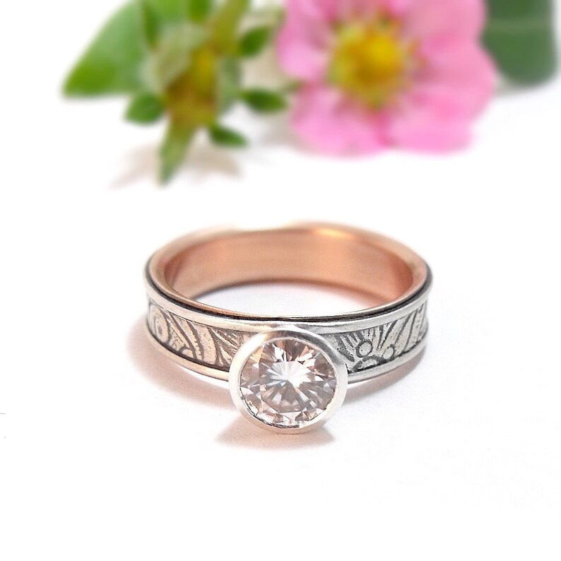 May include: A silver and rose gold ring with a large, round, clear gemstone. The band features an etched design. A pink flower and green leaves are blurred in the background.