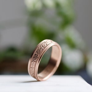 May include: A rose gold wedding band with a delicate floral design engraved around the band.