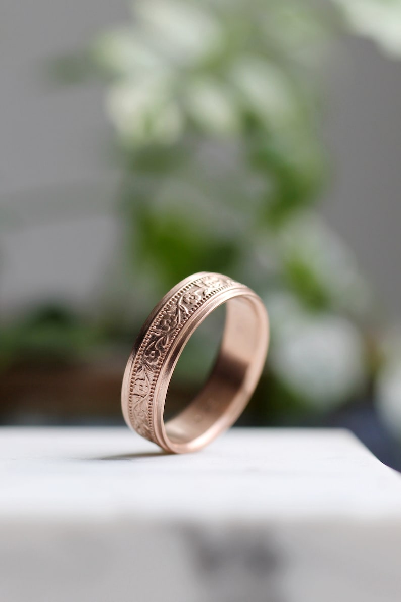 May include: A rose gold wedding band with a delicate floral design engraved around the band.