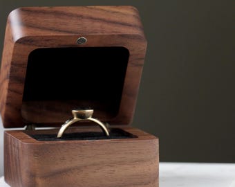 May include: A gold engagement ring with a dark gemstone is displayed in an open, wooden ring box. The box is made of a dark wood with a square shape and a black velvet interior. The ring sits on a white marble surface.
