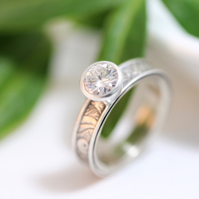 May include: A silver ring with a large, round, clear gemstone. The band features an etched design. The ring is set against a soft, blurred background of white and green.