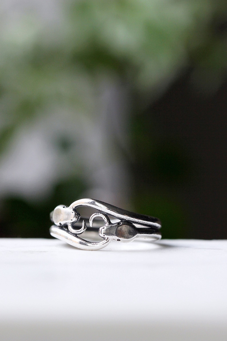 May include: A silver ring with a unique, intricate design featuring two intertwined loops. The ring is made of a smooth, polished metal and has a delicate, feminine feel.