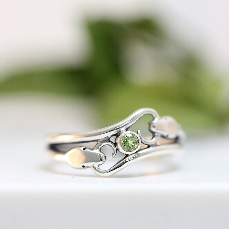 May include: A silver ring with a delicate, swirling design featuring a green gemstone. The ring has a heart-shaped cutout on one side.