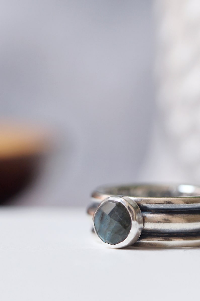 May include: Close-up of a silver ring featuring a faceted, round, blue-grey gemstone. The ring has a textured band with three parallel lines. The background is blurred, with soft neutral tones.