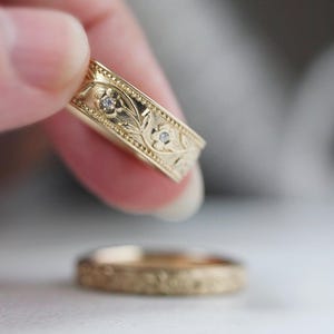 Op de afbeelding: Twee gouden trouwringen met een bloemmotief en kleine diamanten. De ring bovenop heeft een ingewikkelder ontwerp met bladeren en bloemen.