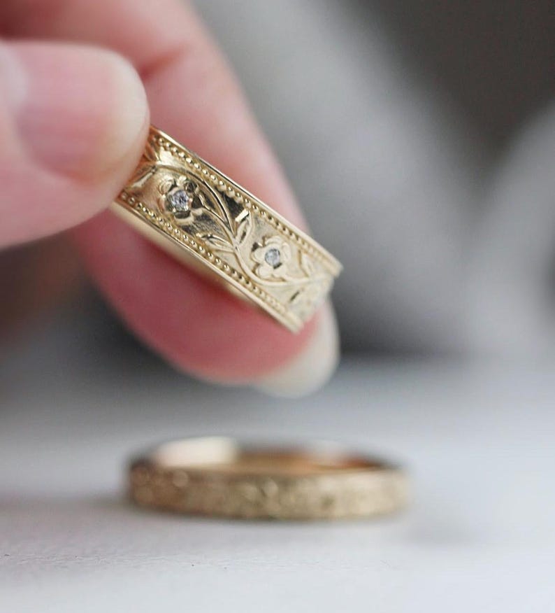Op de afbeelding: Twee gouden trouwringen met een bloemmotief en kleine diamanten. De ring bovenop heeft een ingewikkelder ontwerp met bladeren en bloemen.
