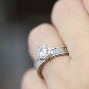 May include: A silver ring with a large, round, clear gemstone set in a bezel. The band features an engraved floral pattern. The ring is on a finger, with a blurred background.