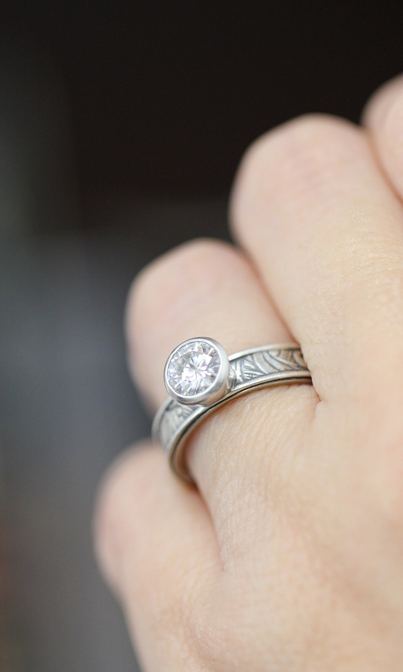 May include: A silver ring with a large, round, clear gemstone set in a bezel. The band features an engraved floral pattern. The ring is on a finger, with a blurred background.