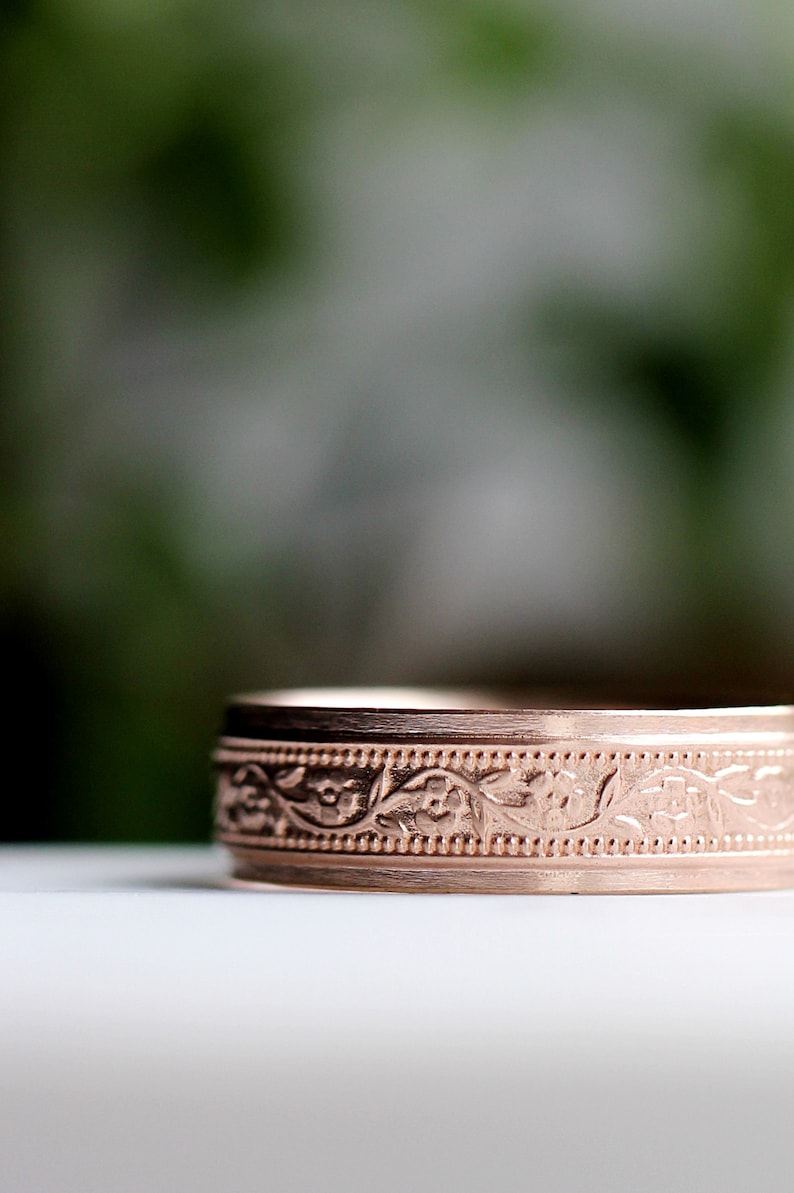 May include: A close-up of a rose gold wedding band with a floral design.