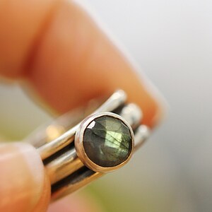 May include: A set of stackable silver rings, each with a polished finish. The top ring features an oval-cut labradorite gemstone, displaying green and blue iridescence. The rings are held by a person's fingers.