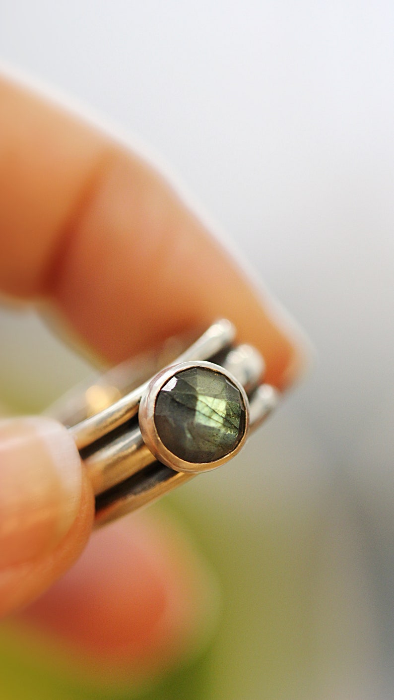 May include: A set of stackable silver rings, each with a polished finish. The top ring features an oval-cut labradorite gemstone, displaying green and blue iridescence. The rings are held by a person's fingers.