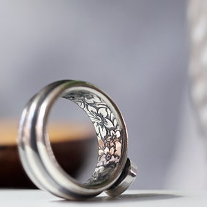 May include: A silver ring with a floral pattern etched inside the band. The ring has a smooth, polished exterior and a small, round stone set on the side. The floral design features delicate flowers and leaves. The ring is likely made of silver.
