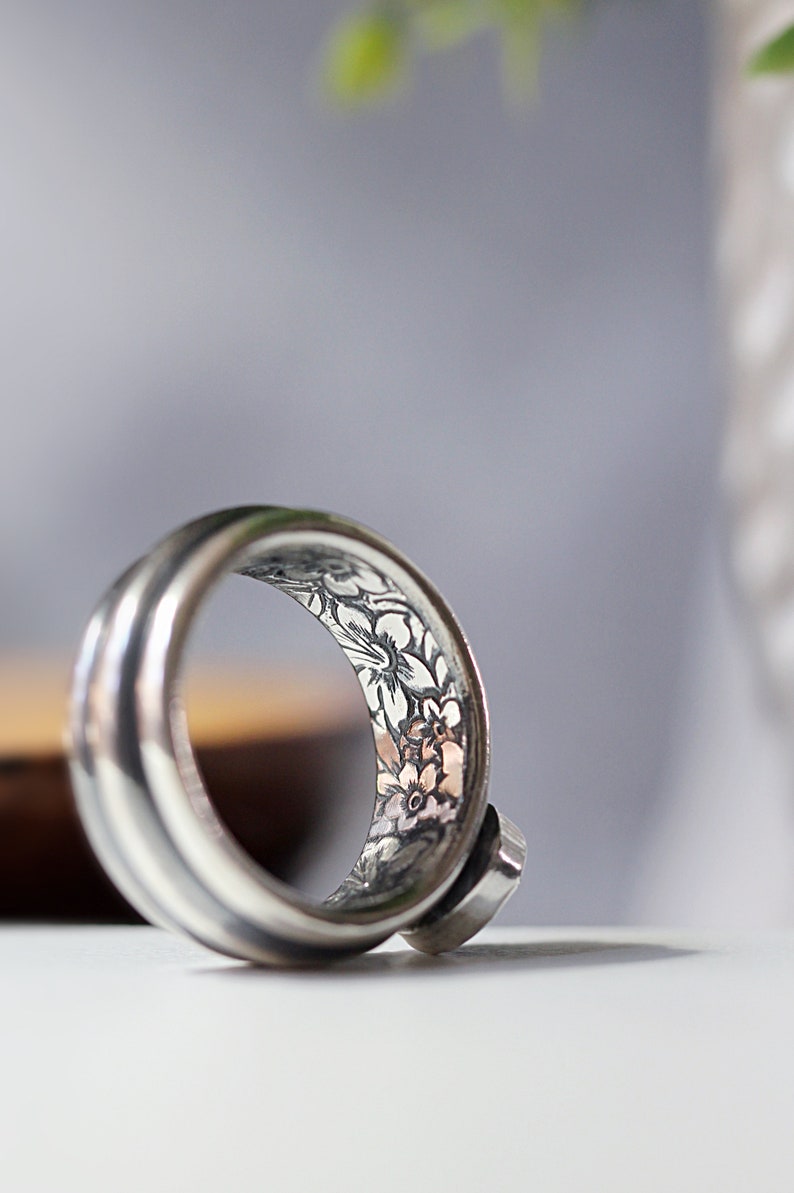 May include: A silver ring with a floral pattern etched inside the band. The ring has a smooth, polished exterior and a small, round stone set on the side. The floral design features delicate flowers and leaves. The ring is likely made of silver.