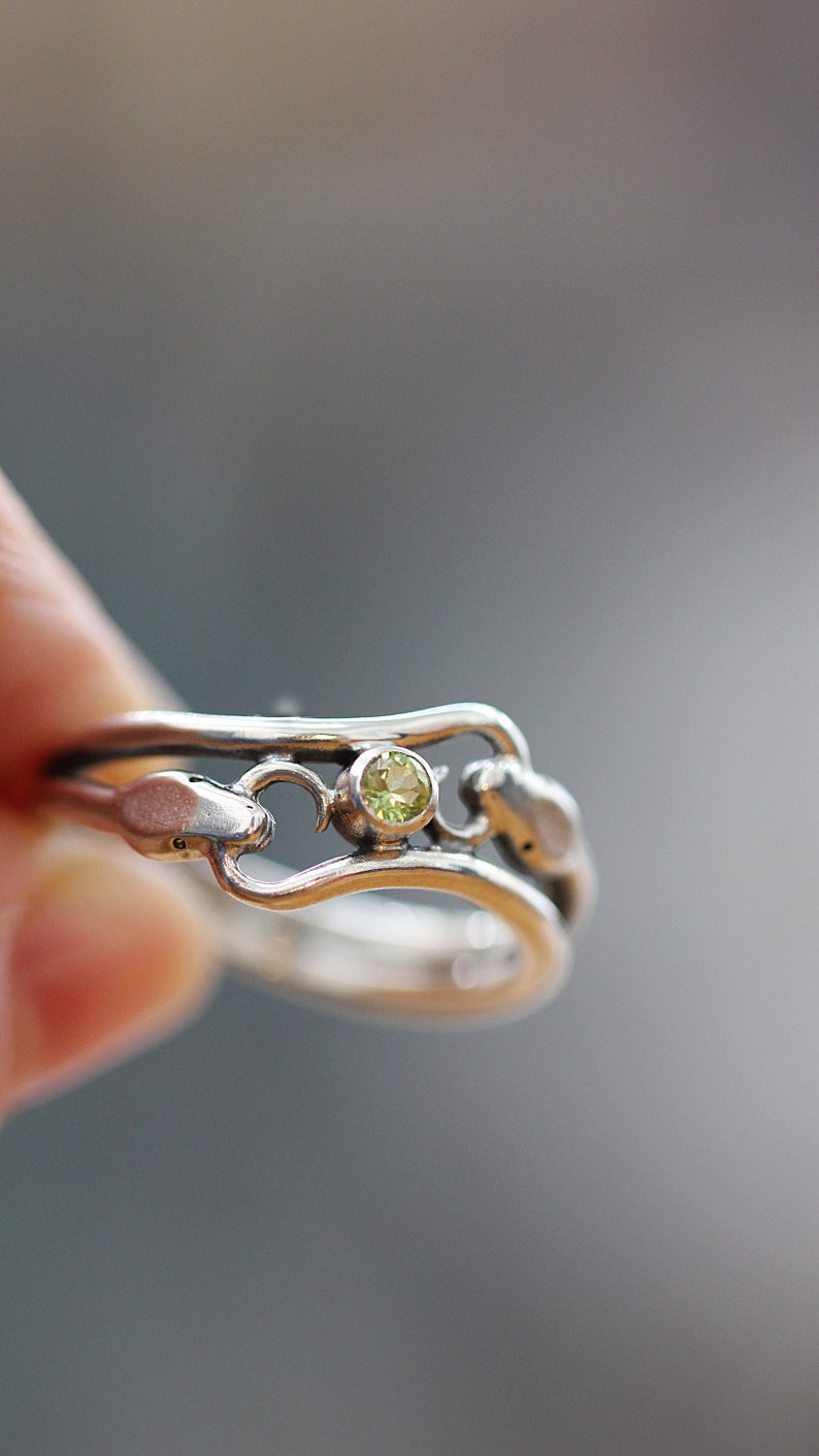 May include: A silver ring with a delicate design featuring a green gemstone. The ring is set with a single, round stone in the center.