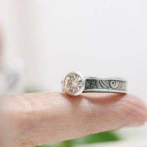 May include: A silver ring with a large, clear, round gemstone. The band features an etched design of swirling patterns. The ring is shown on a finger, with a soft, white background.