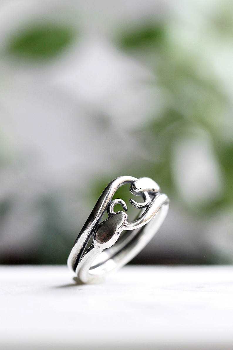May include: A silver ring with a delicate, intricate design. The ring features a wave-like band with a stylized floral motif.