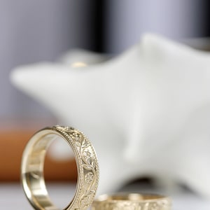 Op de afbeelding: Twee gouden trouwringen met een bloemmotief. De ringen liggen op een wit oppervlak.