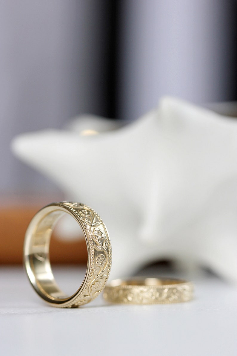 Op de afbeelding: Twee gouden trouwringen met een bloemmotief. De ringen liggen op een wit oppervlak.