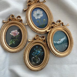 May include: Four oval-shaped gold frames with decorative bow accents at the top. Each frame contains a small floral painting: a pink rose on black, a white flower on blue, a blue flower bouquet in a glass vase, and white daisies on a teal background.