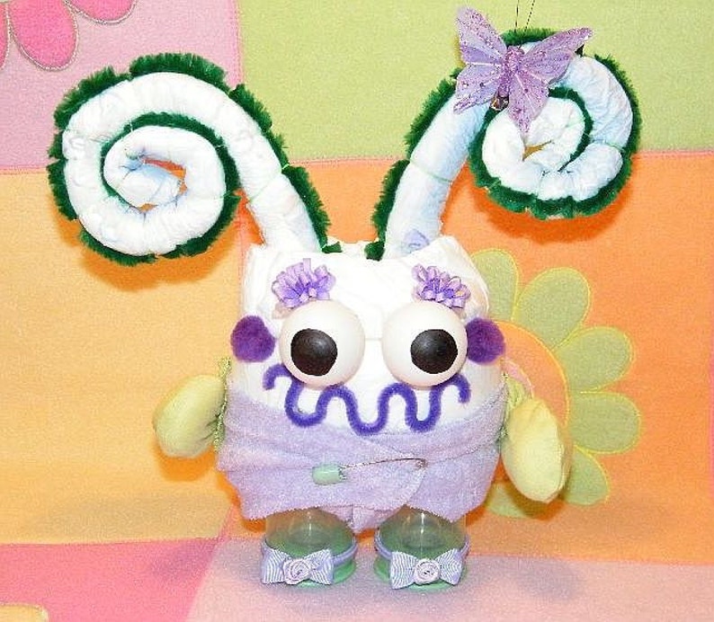 May include: A white diaper cake decorated with green pipe cleaners, purple flowers, and a purple butterfly. The cake is shaped like a monster with two large eyes and a purple mouth.