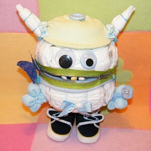 May include: A white diaper cake decorated with blue and green accents. The cake is shaped like a monster with a zipper mouth and large googly eyes. The cake is topped with a blue and white bow.