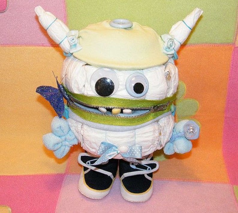 May include: A white diaper cake decorated with blue and green accents. The cake is shaped like a monster with a zipper mouth and large googly eyes. The cake is topped with a blue and white bow.