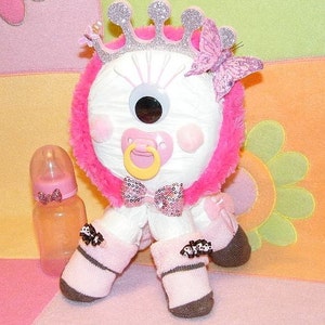 May include: A white stuffed toy with a single large eye, wearing a pink and silver crown, a pink bow, and pink and white booties. The toy is holding a pink and white pacifier.