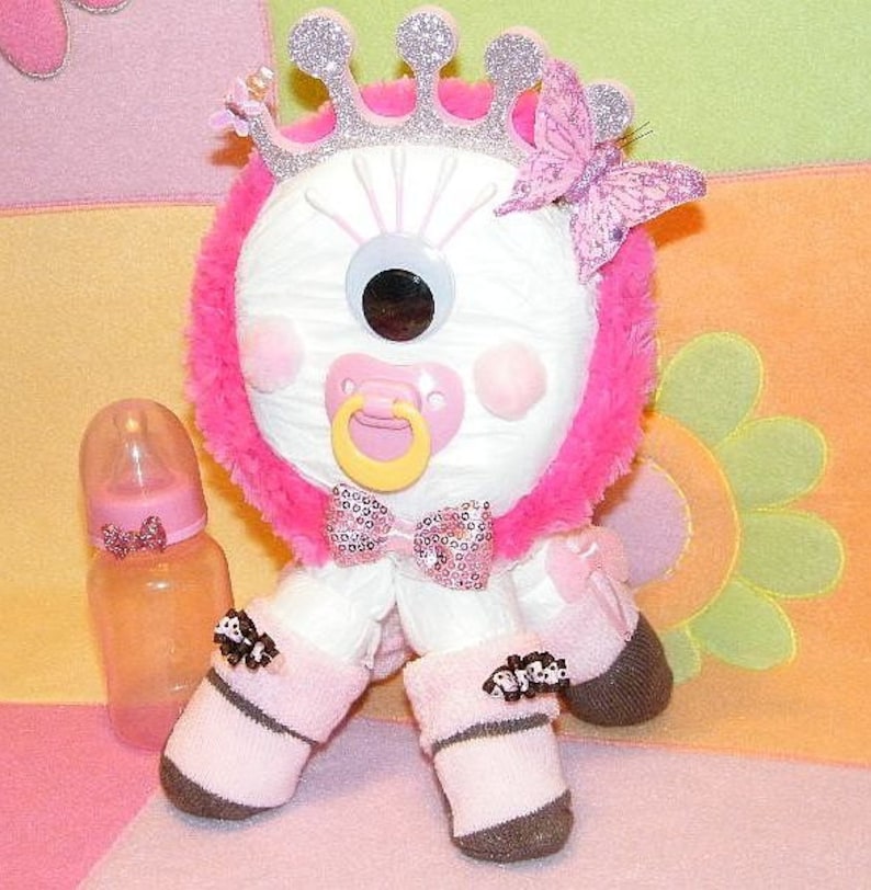 May include: A white stuffed toy with a single large eye, wearing a pink and silver crown, a pink bow, and pink and white booties. The toy is holding a pink and white pacifier.