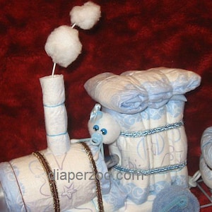 Puede incluir: Un pastel de pa&ntilde;ales azul y blanco con forma de tren con un animal de peluche azul y blanco en la parte superior. El tren tiene una chimenea de algod&oacute;n blanco y una cinta marr&oacute;n alrededor. El pastel est&aacute; decorado con cinta azul y blanca y una tela estampada azul y blanca.