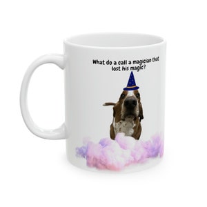 May include: White ceramic mug with a brown and white basset hound wearing a blue and gold wizard hat. The mug has the text "What do you call a magician that lost his magic?" printed on it.