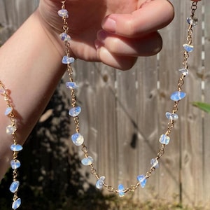 May include: A delicate necklace featuring small, iridescent, pale blue-white opal stones strung on a gold-colored chain. The necklace is shown in a close-up view, highlighting the details of the gemstones and the chain's design. The jewelry is displayed against a blurred background.