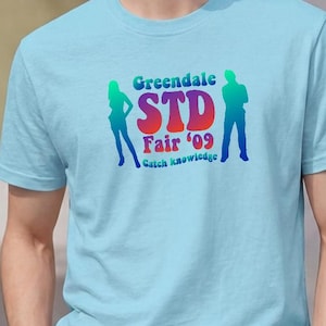 May include: A light blue short-sleeved t-shirt featuring a colorful graphic print on the chest. The design includes the text Greendale STD Fair '09 Catch knowledge in a rainbow gradient, flanked by two dark blue silhouettes of standing figures.