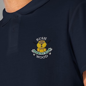 May include: A navy blue short-sleeved polo shirt featuring an embroidered graphic on the left chest. The graphic depicts a brown gopher with the text "BUSH COUNTRY CLUB WOOD" and two crossed golf clubs behind the gopher on a light banner.