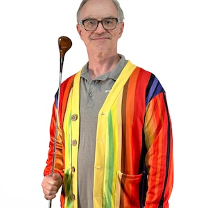 May include: A man wearing a colorful striped cardigan with a yellow background and a grey polo shirt underneath. He is holding a wooden-handled golf club.