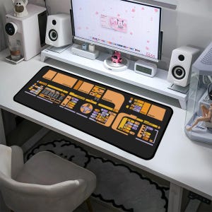 Pode incluir: Um tapete de mesa preto e laranja com um design detalhado de um painel de controle de nave espacial. O tapete está em uma mesa branca com um computador, alto-falantes e outros materiais de escritório.