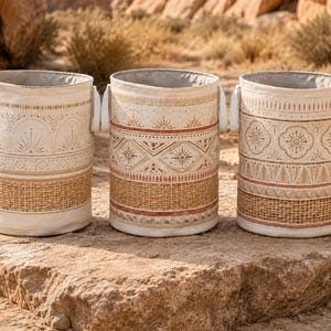 Peut inclure: Trois paniers de rangement cylindriques en tissu blanc cassé et en fibres naturelles tressées. Chaque panier présente des motifs géométriques imprimés distincts dans des tons marron et terre cuite, avec des anses latérales blanches.
