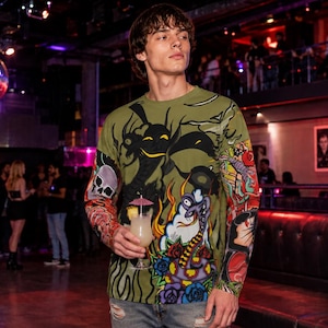 Peut inclure: Un t-shirt à manches longues vert olive arborant un imprimé graphique complexe comprenant un crâne, des silhouettes stylisées et des éléments floraux aux couleurs vives. Le motif couvre le devant et les manches, porté avec un jean bleu clair usé.