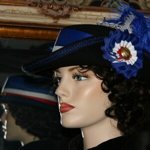 May include: A black hat with blue ribbon and feather detailing. The hat has a blue, white and red rosette with a gold button. The hat is displayed on a mannequin.